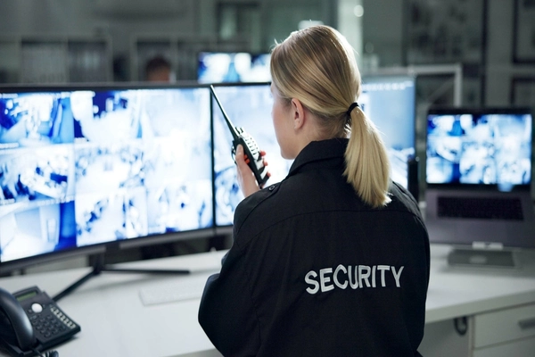eine Sicherheitsdienstmitarbeiterin sitzt vor mehrere Monitoren und hat ein Funkgerät in der Hand